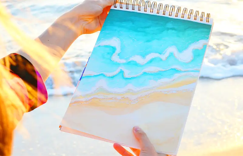 L'artiste peintre Feel tient dans ses mains son oeuvre Ecume représentant la plage et les vagues qu'elle positionne devant la plage et les vagues de la mer méditerrannée. Affiche de Expositions Toulon. Live painting hyères